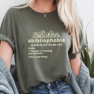 Pu&ograve; includere: T-shirt verde oliva con la parola "abibliophobia" e la sua pronuncia tra parentesi. Sotto, la definizione della parola: "La paura di rimanere senza libri. &Egrave; una cosa reale."