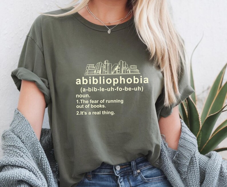 Pu&ograve; includere: T-shirt verde oliva con la parola "abibliophobia" e la sua pronuncia tra parentesi. Sotto, la definizione della parola: "La paura di rimanere senza libri. &Egrave; una cosa reale."