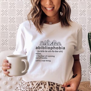 Pu&ograve; includere: Maglietta bianca con la parola "abibliophobia" stampata in nero, insieme alla sua pronuncia e definizione. La maglietta presenta una grafica di libri impilati. Una persona tiene in mano una tazza bianca.