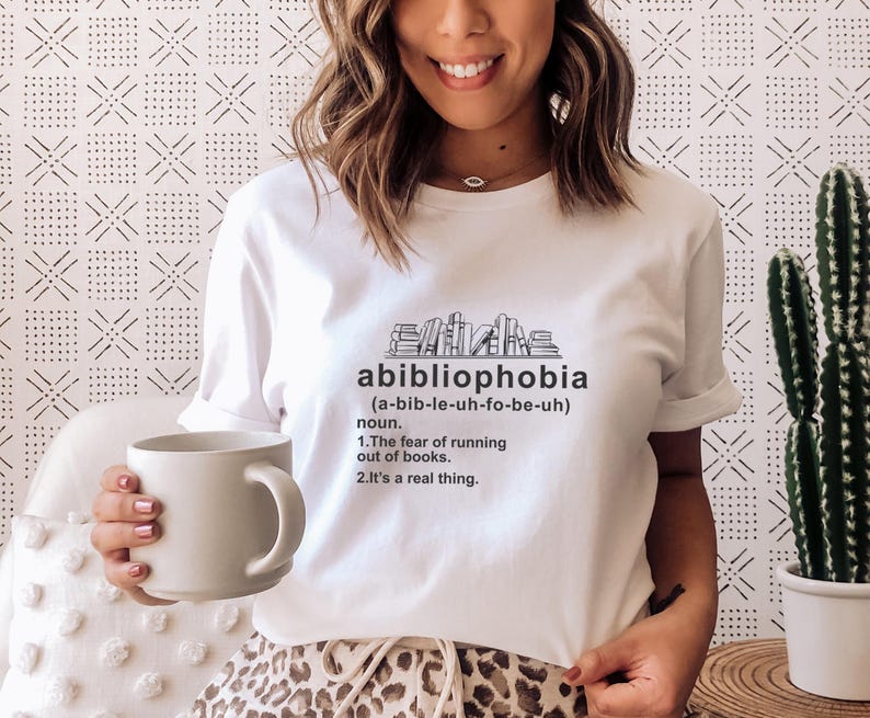 Pu&ograve; includere: Maglietta bianca con la parola "abibliophobia" stampata in nero, insieme alla sua pronuncia e definizione. La maglietta presenta una grafica di libri impilati. Una persona tiene in mano una tazza bianca.