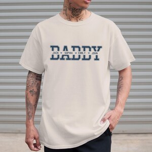 May include: A white t-shirt with a navy blue graphic that says "Daddy" with the names "Jack", "Sophia", "Emily", and "John" underneath.