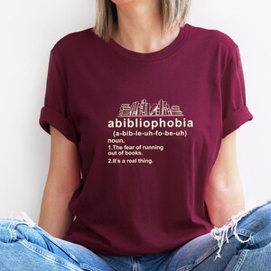 Pu&ograve; includere: Una maglietta bordeaux con la parola "abibliophobia" e la sua definizione stampata in bianco. La definizione include la pronuncia, la parte del discorso e il significato: "la paura di rimanere senza libri."