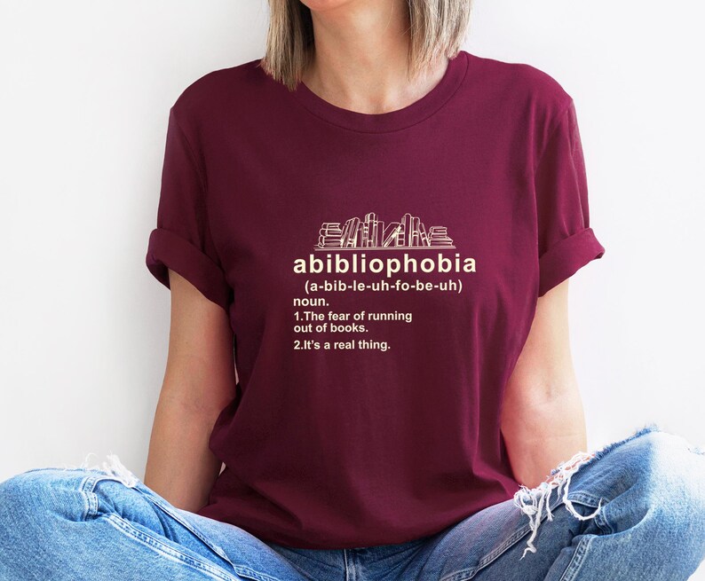 Pu&ograve; includere: Una maglietta bordeaux con la parola "abibliophobia" e la sua definizione stampata in bianco. La definizione include la pronuncia, la parte del discorso e il significato: "la paura di rimanere senza libri."
