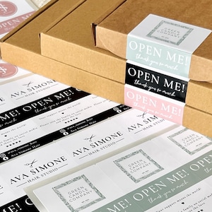 May include: A close-up of several small business stickers with the text "Open Me! Thank you so much" printed on them. The stickers are in various colours, including black, white, pink, and green. The stickers are arranged in a random pattern on a white surface.