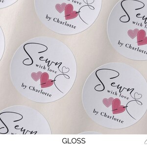 K&ouml;nnte beinhalten: Runde wei&szlig;e Aufkleber mit dem Text "Sewn with love by Charlotte" und einem rosa Herz mit einer Nadel und einem Faden, der durch das Herz geht.