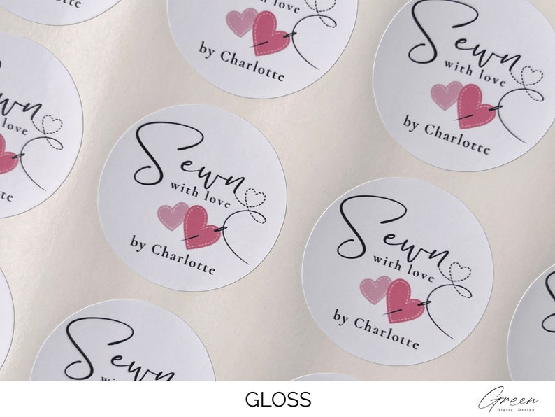 K&ouml;nnte beinhalten: Runde wei&szlig;e Aufkleber mit dem Text "Sewn with love by Charlotte" und einem rosa Herz mit einer Nadel und einem Faden, der durch das Herz geht.