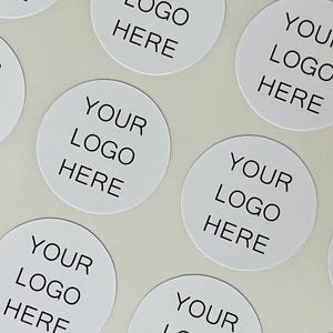 May include: A collection of circular white stickers arranged in a pattern. Each sticker features the black text "YOUR LOGO HERE" printed in a clean, sans-serif font, centered on the white surface of the individual round adhesive labels.