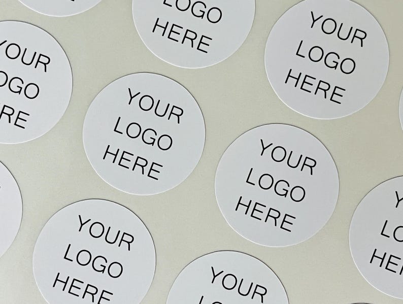 May include: A collection of circular white stickers arranged in a pattern. Each sticker features the black text "YOUR LOGO HERE" printed in a clean, sans-serif font, centered on the white surface of the individual round adhesive labels.