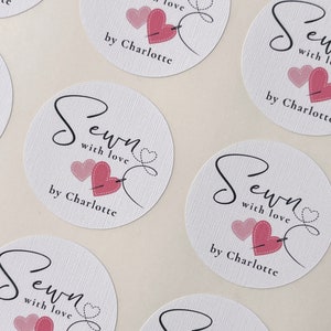 K&ouml;nnte beinhalten: Runde wei&szlig;e Aufkleber mit rosa Herzen und einem Nadel- und Fadenmuster. Der Text lautet "Sewn with love by Charlotte".