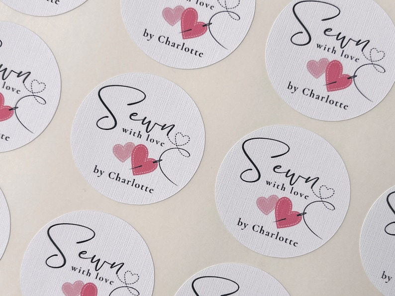 K&ouml;nnte beinhalten: Runde wei&szlig;e Aufkleber mit rosa Herzen und einem Nadel- und Fadenmuster. Der Text lautet "Sewn with love by Charlotte".