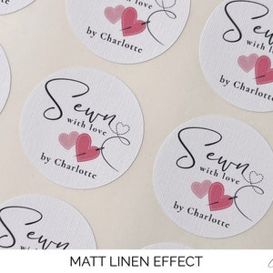 K&ouml;nnte beinhalten: Runde wei&szlig;e Aufkleber mit einem rosa Herz- und Nadelmotiv. Der Text "Sewn with love by Charlotte" ist auf den Aufklebern gedruckt.