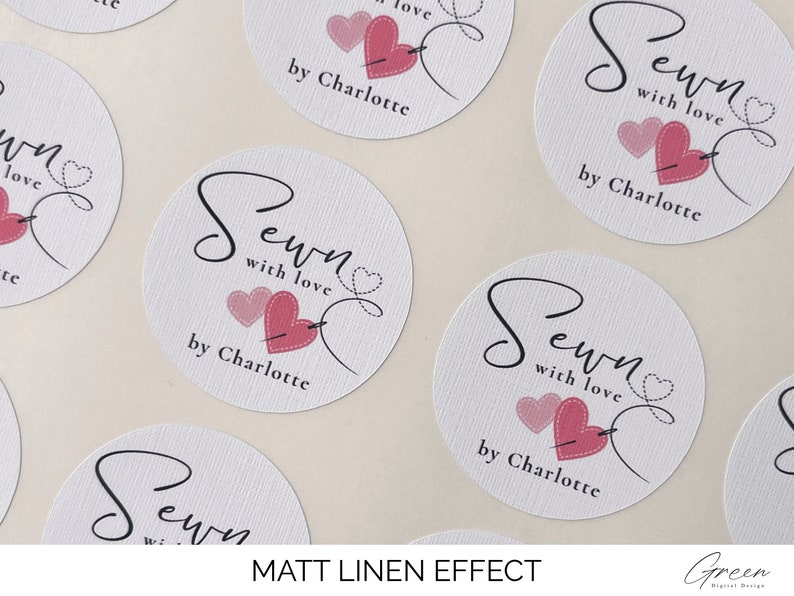 K&ouml;nnte beinhalten: Runde wei&szlig;e Aufkleber mit einem rosa Herz- und Nadelmotiv. Der Text "Sewn with love by Charlotte" ist auf den Aufklebern gedruckt.