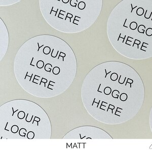 May include: A collection of circular white stickers arranged in a pattern. Each sticker features the black printed text "YOUR LOGO HERE" centered in a clean, sans-serif font. The stickers have a matte finish and are presented on a light background.