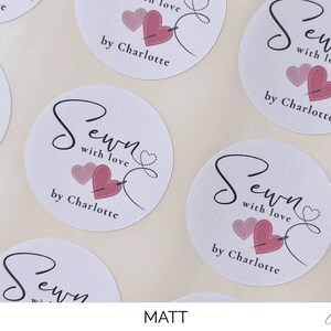 K&ouml;nnte beinhalten: Runde wei&szlig;e Aufkleber mit dem Text "Sewn with love by Charlotte" in schwarzer Schrift. Zwei rosa Herzen mit einer Nadel und Faden sind neben dem Text abgebildet.