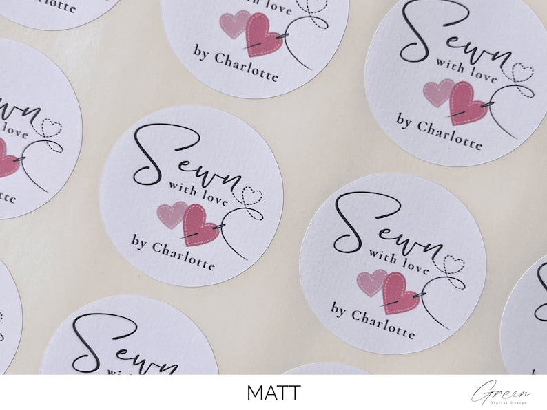 K&ouml;nnte beinhalten: Runde wei&szlig;e Aufkleber mit dem Text "Sewn with love by Charlotte" in schwarzer Schrift. Zwei rosa Herzen mit einer Nadel und Faden sind neben dem Text abgebildet.