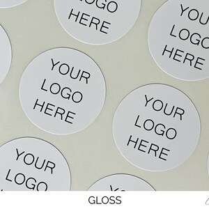 May include: A collection of circular white stickers featuring the black text YOUR LOGO HERE printed in the center. The stickers have a smooth, reflective gloss finish and are arranged in a repeating pattern across the frame for branding purposes.