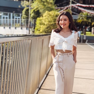 Puede incluir: Una mujer con un top blanco con encaje, pantalones beige de cintura alta y cintur&oacute;n de cadena dorada. Camina por un puente de madera. El conjunto se complementa con sandalias. El fondo es soleado.