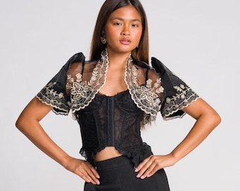 May include: A black lace corset top with a front hook-and-eye closure, paired with a sheer black bolero featuring intricate gold floral embroidery on the sleeves and collar, worn with high-waisted black wide-leg trousers and black sandals.