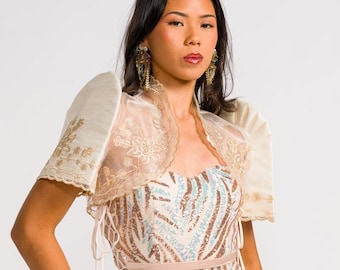 Könnte beinhalten: Ein bodenlanges, trägerloses Kleid mit einem Paillettenmuster aus bronzefarbenen und hellblauen Blättern auf cremefarbenem Grund, kombiniert mit einem transparenten cremefarbenen Bolero mit strukturierten Schmetterlingsärmeln und Goldstickerei.