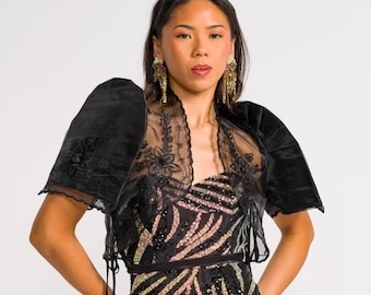 May include: A floor-length strapless gown featuring a black base with iridescent, multi-colored sequined zebra-like patterns. The gown is paired with a sheer black bolero jacket that has short, puffed sleeves and intricate black floral embroidery.