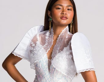May include: A white floor-length dress featuring iridescent sequins in a swirling pattern, paired with a sheer white bolero jacket. The bolero has structured butterfly sleeves and intricate floral embroidery along the lapels and edges.