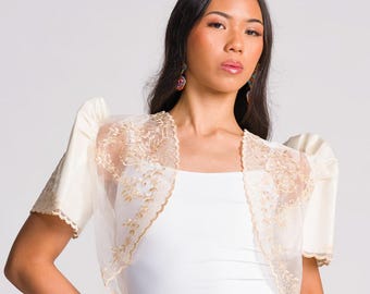 May include: A cream-colored bolero jacket featuring sheer organza panels with intricate gold floral embroidery, puffed sleeves, and scalloped edges. The garment is worn over a plain white tank top and a light beige wrap skirt with a side tie detail.
