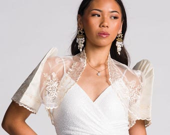 May include: A white sequined wrap dress paired with a sheer cream-colored bolero jacket featuring intricate floral embroidery, scalloped lace edges, and structured butterfly sleeves. The bolero is worn over a sleeveless dress with a V-neckline.