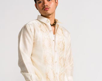 May include: A long-sleeved, off-white button-down shirt featuring a semi-sheer fabric with intricate, symmetrical gold-toned embroidery panels running vertically along the front placket, paired with light beige trousers.