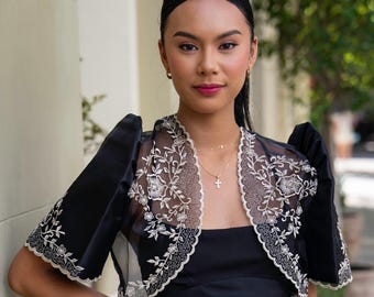 May include: A black bolero jacket featuring sheer fabric with intricate white floral embroidery along the front edges and puffed sleeves, worn over a simple black dress with a square neckline and thin shoulder straps.
