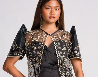 May include: A black bolero jacket featuring sheer panels with intricate gold floral embroidery and structured butterfly sleeves, worn over a black sleeveless jumpsuit with a wrap-style bodice and a single pearl-like button closure at the neck.