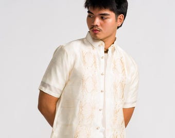 May include: A short-sleeved, off-white button-down shirt featuring a pointed collar and intricate, light brown vertical embroidery panels along the front placket. The garment is worn over a plain white undershirt and paired with light beige trousers.