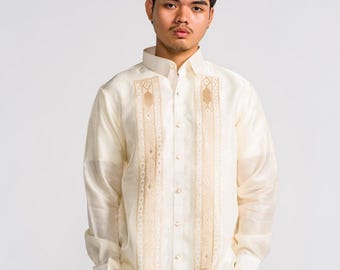 May include: A long-sleeved, off-white Barong Tagalog shirt featuring a structured collar, a button-down front, and two vertical panels of intricate, light brown geometric embroidery running down the chest. The fabric has a sheer, crisp texture.