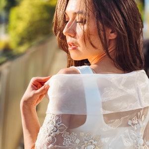 Puede incluir: Un chal blanco transparente bordado con dise&ntilde;os florales y de hojas. El chal se lleva sobre una parte superior blanca. El bordado presenta detalles florales en relieve. La imagen est&aacute; tomada con luz natural, con un enfoque suave en el sujeto.