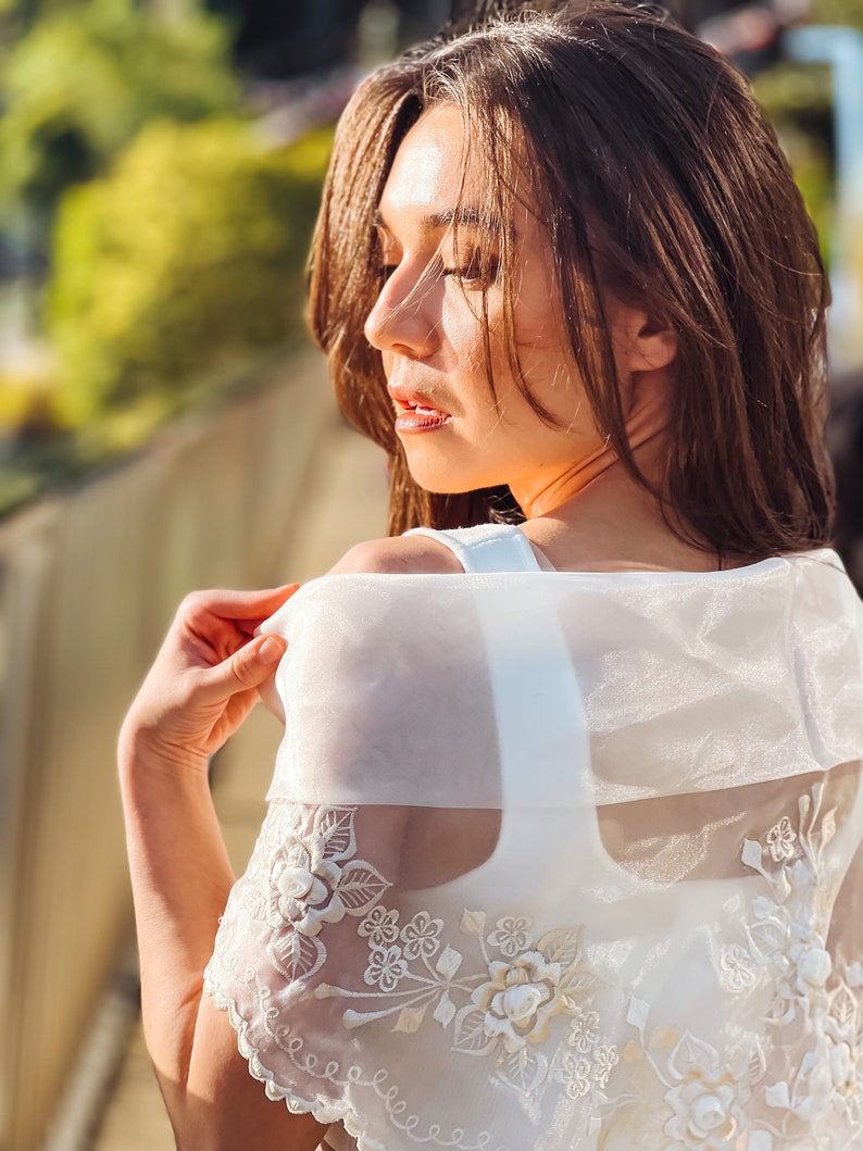 Puede incluir: Un chal blanco transparente bordado con dise&ntilde;os florales y de hojas. El chal se lleva sobre una parte superior blanca. El bordado presenta detalles florales en relieve. La imagen est&aacute; tomada con luz natural, con un enfoque suave en el sujeto.