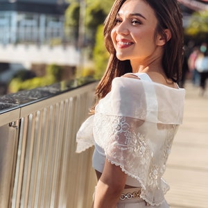 Puede incluir: Una mujer con un top blanco con hombros descubiertos con detalles de encaje floral y un cintur&oacute;n dorado. Sonr&iacute;e y mira hacia un lado. El fondo incluye una pasarela de madera y vegetaci&oacute;n.