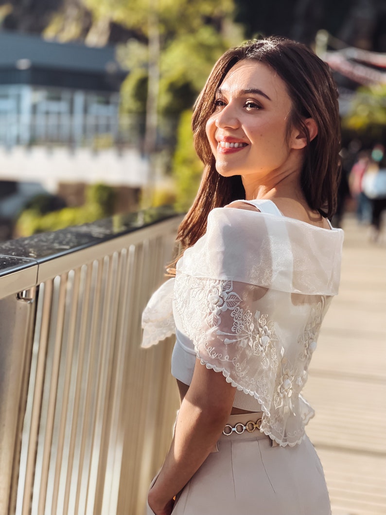 Puede incluir: Una mujer con un top blanco con hombros descubiertos con detalles de encaje floral y un cintur&oacute;n dorado. Sonr&iacute;e y mira hacia un lado. El fondo incluye una pasarela de madera y vegetaci&oacute;n.
