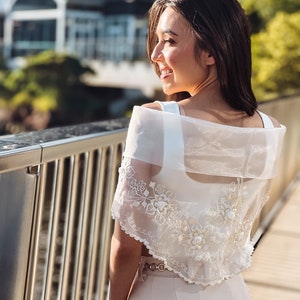 Puede incluir: Bolero crema bordado con detalles florales. Se lleva sobre un top blanco y se combina con pantalones a juego. El conjunto se complementa con un cintur&oacute;n dorado. El fondo muestra un puente y un edificio.