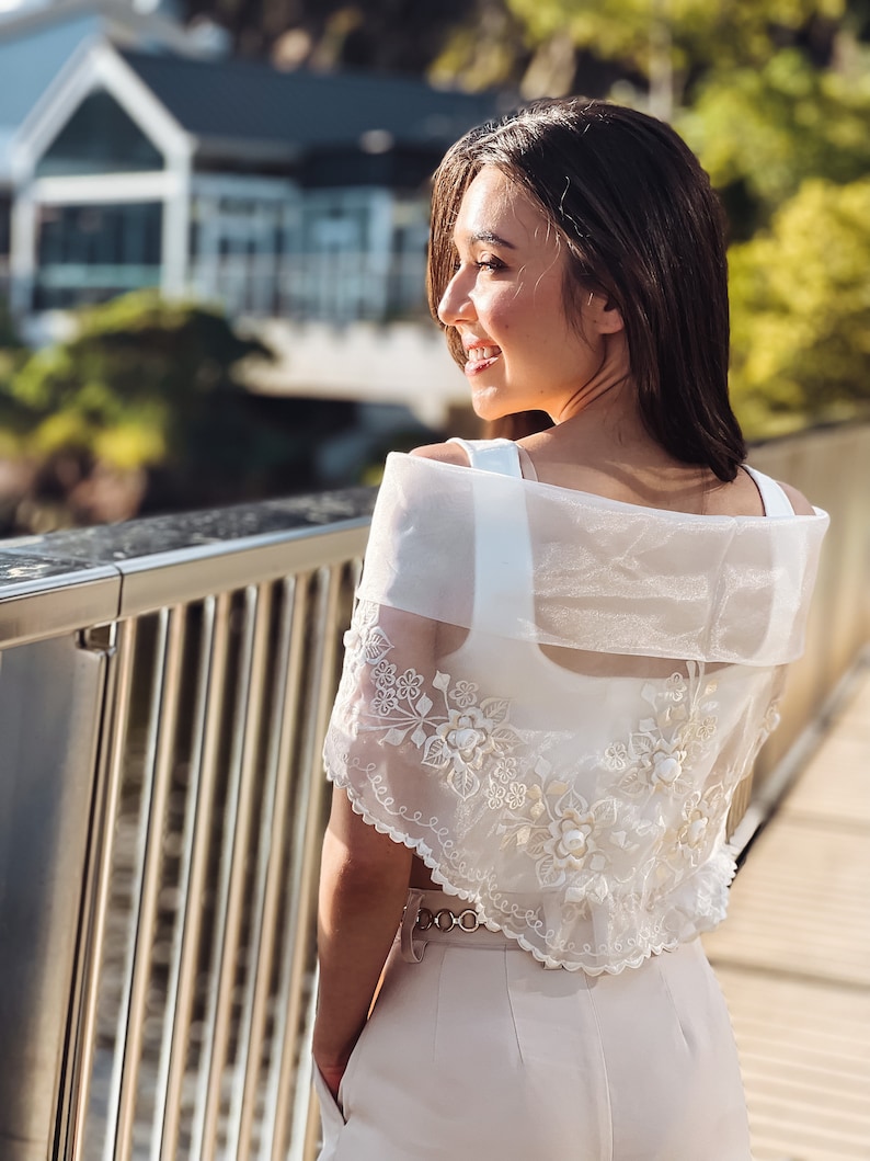 Puede incluir: Bolero crema bordado con detalles florales. Se lleva sobre un top blanco y se combina con pantalones a juego. El conjunto se complementa con un cintur&oacute;n dorado. El fondo muestra un puente y un edificio.