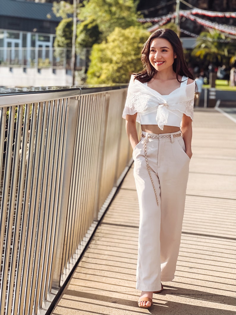 Puede incluir: Una mujer con un top blanco con encaje, pantalones beige de cintura alta y cintur&oacute;n de cadena dorada. Camina por un puente de madera. El conjunto se complementa con sandalias. El fondo es soleado.