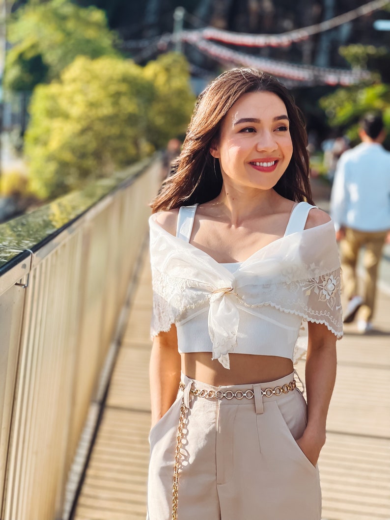 Puede incluir: Una mujer con un top corto blanco, pantalones beige y un cintur&oacute;n de cadena dorada. Tambi&eacute;n lleva un chal de encaje blanco transparente. El fondo incluye un puente de madera, vegetaci&oacute;n y una persona con una camisa blanca.