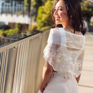 Puede incluir: Bolero blanco transparente bordado con detalles florales. El bolero se lleva sobre un top blanco y se combina con pantalones de color claro. Un cintur&oacute;n plateado ci&ntilde;e la cintura. El fondo presenta un puente y vegetaci&oacute;n.