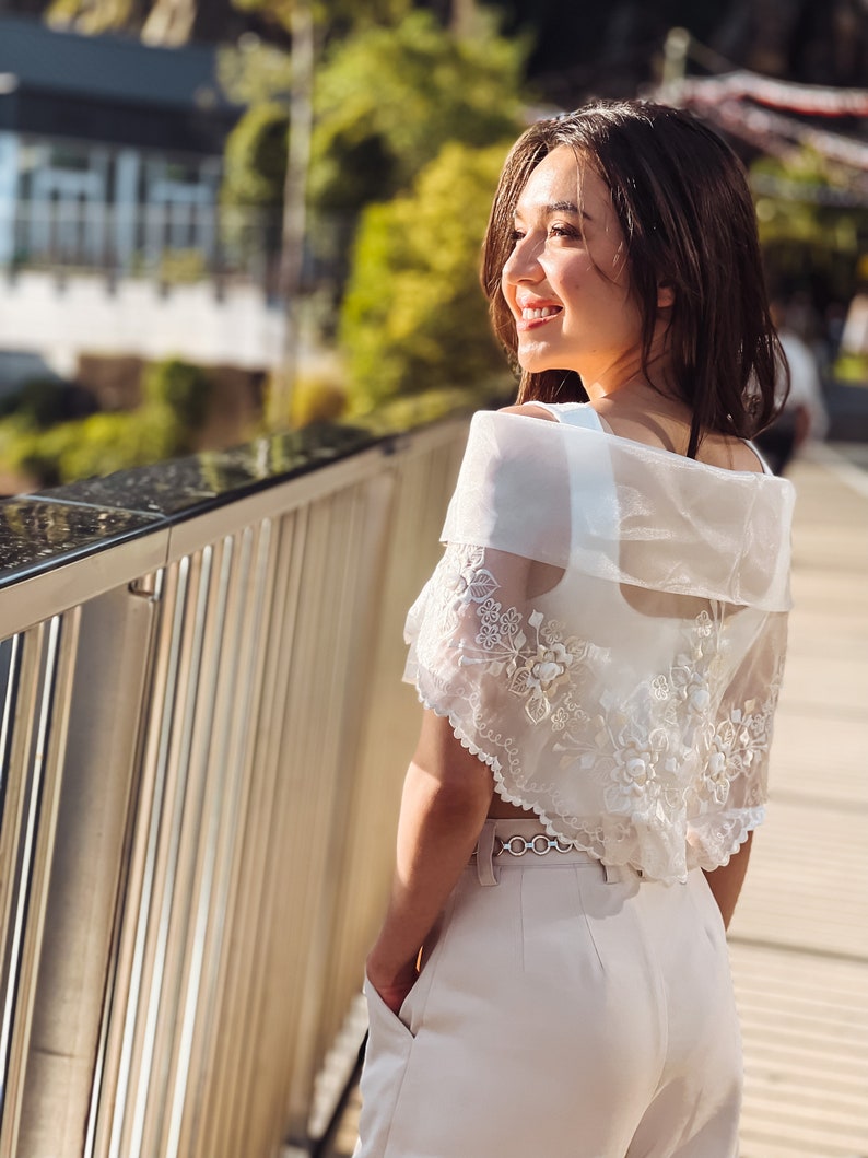 Puede incluir: Bolero blanco transparente bordado con detalles florales. El bolero se lleva sobre un top blanco y se combina con pantalones de color claro. Un cintur&oacute;n plateado ci&ntilde;e la cintura. El fondo presenta un puente y vegetaci&oacute;n.