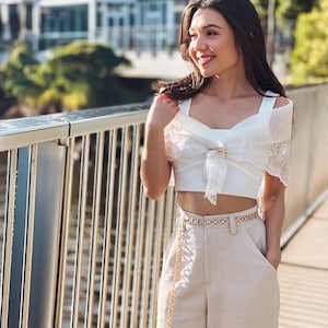 Puede incluir: Un top corto blanco con una superposici&oacute;n transparente con ribete de encaje y un cintur&oacute;n de cadena dorado. El atuendo se combina con pantalones de cintura alta beige. El look general es elegante y con estilo, adecuado para un d&iacute;a de verano.