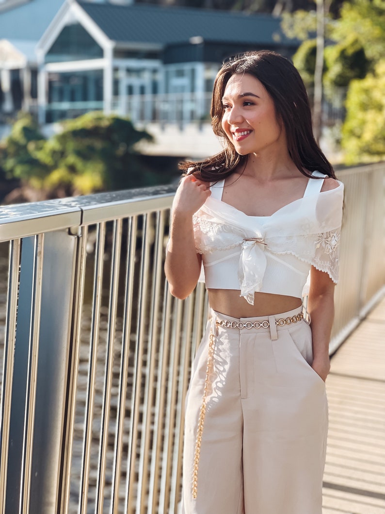 Puede incluir: Un top corto blanco con una superposici&oacute;n transparente con ribete de encaje y un cintur&oacute;n de cadena dorado. El atuendo se combina con pantalones de cintura alta beige. El look general es elegante y con estilo, adecuado para un d&iacute;a de verano.