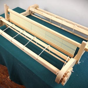 May include: A light-coloured wooden rigid heddle loom featuring a central reed with vertical slots, two parallel warp beams with tensioning knobs on the sides, and white cord ties connecting the beams to the frame for weaving textile projects.
