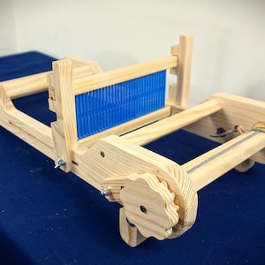 May include: A light-coloured wooden weaving loom featuring a bright blue plastic heddle reed. The device includes two side-mounted adjustment knobs with a gear-like shape, horizontal wooden beams, and metal hardware components for tension control.