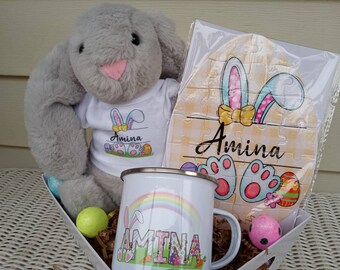 May include: A gray plush bunny wearing a white t-shirt with the name "Amina" printed on it. The bunny is in a basket with a white mug that says "Amina" and a puzzle with the name "Amina" on it. The puzzle is shaped like an Easter egg with bunny ears.