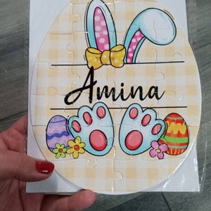 May include: A personalized Easter egg puzzle with a yellow, white, and blue bunny design. The puzzle features a yellow bow, pink and white polka dots, and the name "Amina" written in black. The puzzle is surrounded by a yellow and white checkered background.