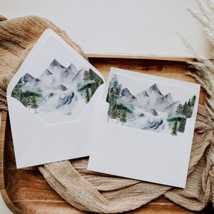 May include: Two white envelopes featuring a watercolour design of mountains and trees. The envelopes are displayed on a wooden tray with a brown fabric underneath.