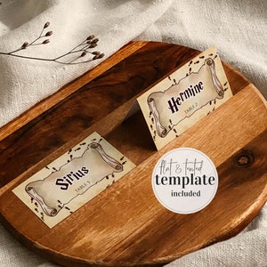 Puede incluir: Dos tarjetas de mesa imprimibles con temática de Harry Potter con un diseño de papel pergamino. Las tarjetas presentan los nombres "Sirius" y "Hermione" con los números de mesa "3" y "2" respectivamente. El texto "plantilla plana y en forma de tienda incluida" está escrito en un círculo blanco.